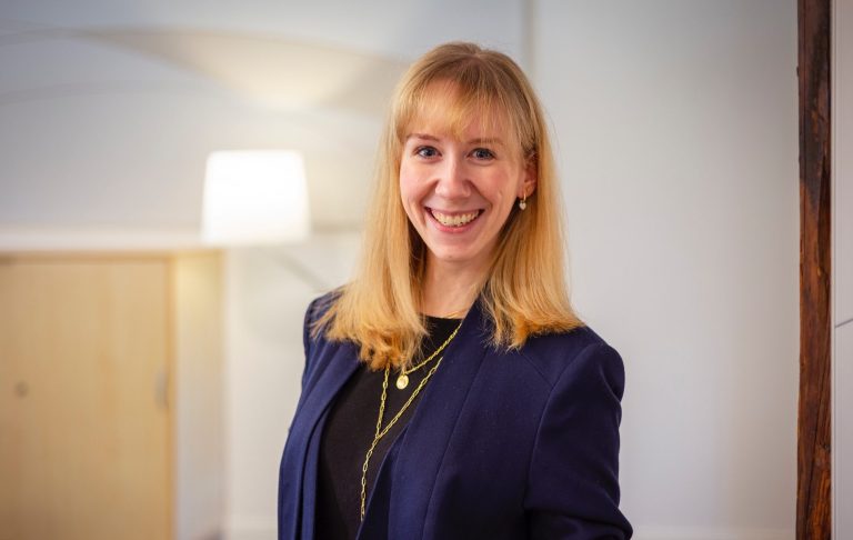 Lächelnde Frau mit blonden Haaren in einem blauen Blazer vor hellem Hintergrund.