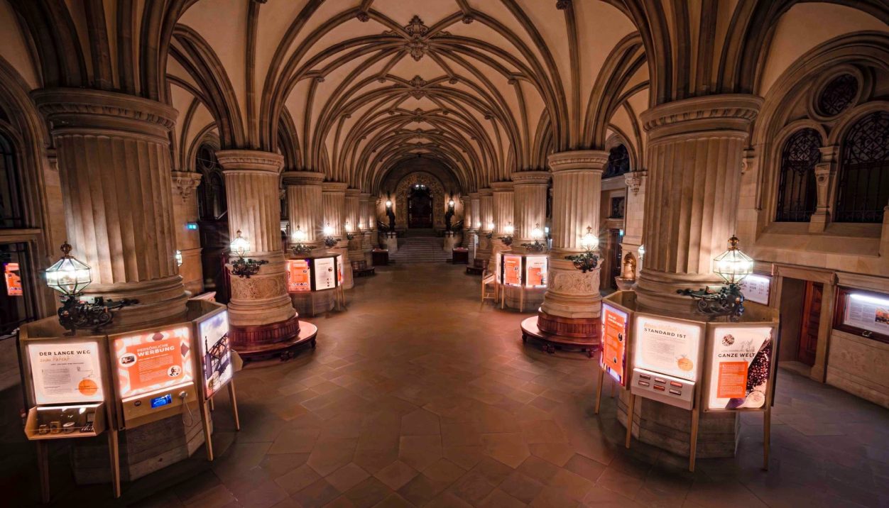 Blick in einen langen, gewölbten Gang mit Säulen und Ausstellungsstafeln.