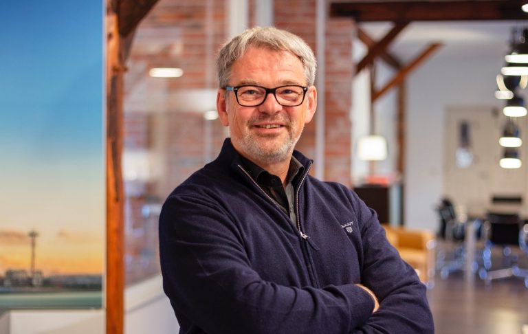 Ein Mann mit Brille, lächelnd, in einem modernen Büroumgebung.
