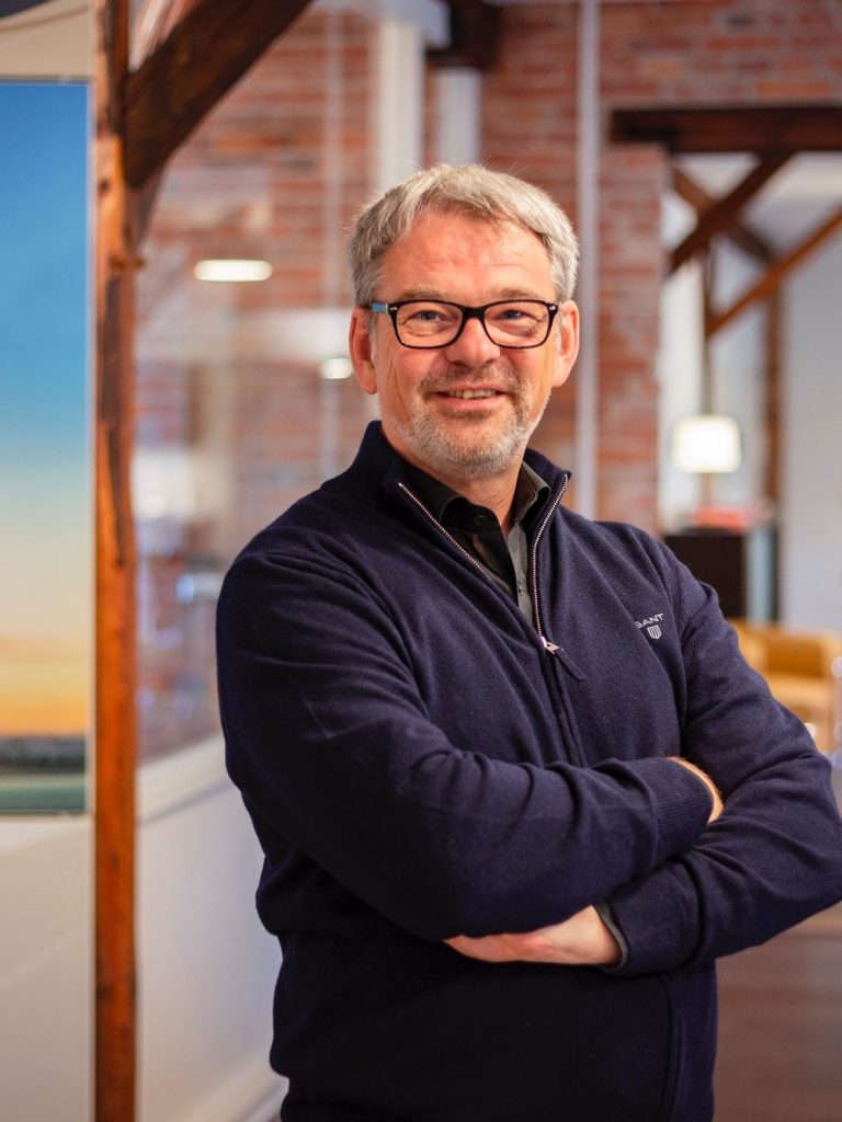 Mann mit Brille und grauem Haar, lächelt in einer modernen Büroumgebung.