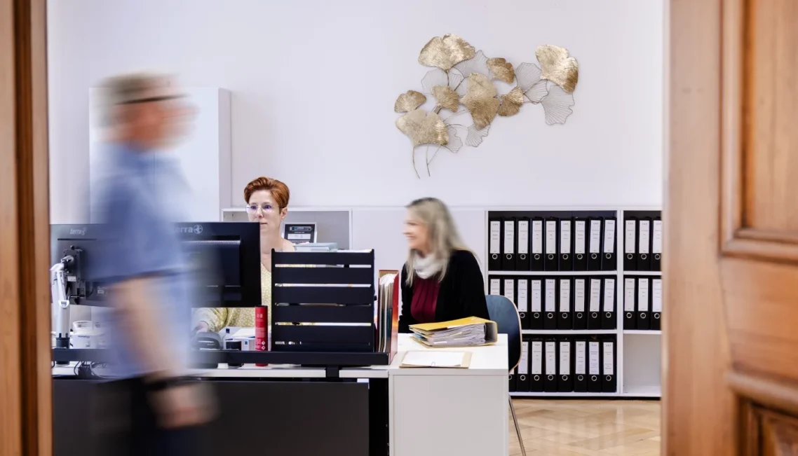 Zwei Frauen im Büro sitzen an einem Schreibtisch, eine Person geht vorbei. Aktenschränke im Hintergrund.